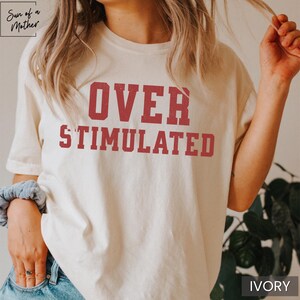 May include: A white t-shirt with the text "OVER STIMULATED" printed in red on the front. The shirt is being worn by a person with long brown hair.