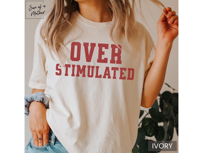May include: A white t-shirt with the text "OVER STIMULATED" printed in red on the front. The shirt is being worn by a person with long brown hair.
