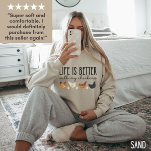 May include: A woman wearing a light beige sweatshirt with the text "LIFE IS BETTER with my chickens" and a row of five chickens printed on the front. The woman is sitting on a carpeted floor with her legs crossed.