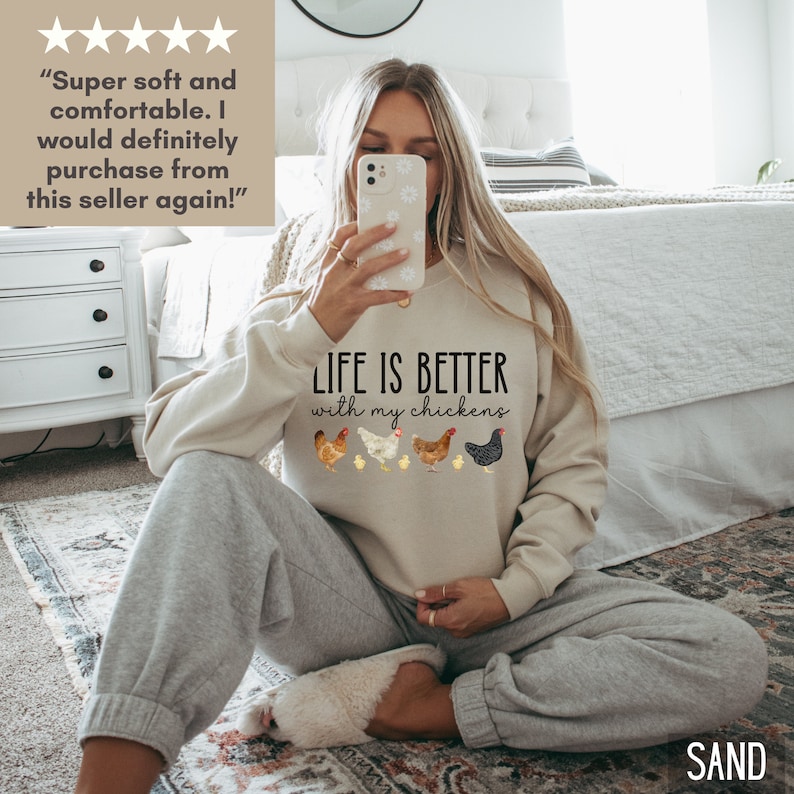 May include: A woman wearing a light beige sweatshirt with the text "LIFE IS BETTER with my chickens" and a row of five chickens printed on the front. The woman is sitting on a carpeted floor with her legs crossed.