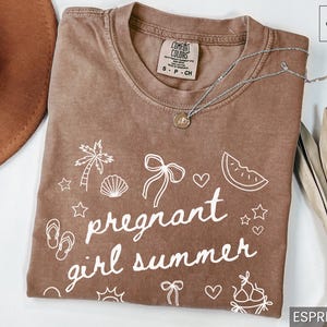 May include: Espresso-colored t-shirt with the words "pregnant girl summer" in white script, along with beach-themed doodles. A silver necklace with a pendant rests on the shirt. A brown hat and dried leaves are in the background.