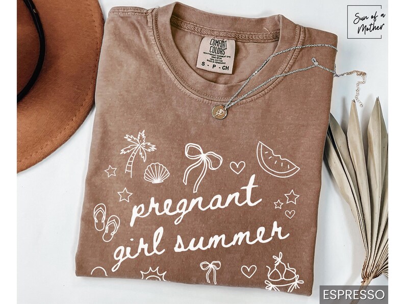 May include: Espresso-colored t-shirt with the words "pregnant girl summer" in white script, along with beach-themed doodles. A silver necklace with a pendant rests on the shirt. A brown hat and dried leaves are in the background.