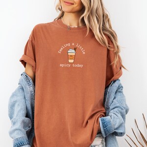 May include: Rust-colored t-shirt with the text "feeling a little spicy today" and a graphic of a pumpkin spice latte. The shirt is paired with a denim jacket. The design is centered on the front of the shirt.