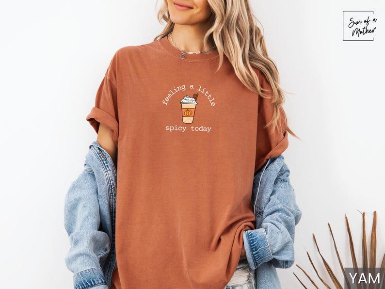 May include: Rust-colored t-shirt with the text "feeling a little spicy today" and a graphic of a pumpkin spice latte. The shirt is paired with a denim jacket. The design is centered on the front of the shirt.