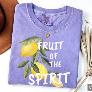 Op de afbeelding: Een paars T-shirt met de tekst "Fruit of the Spirit" in witte letters. Het shirt heeft een aquarel-illustratie van citroenen aan een tak.