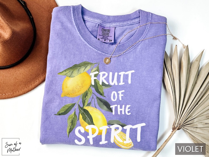 Op de afbeelding: Een paars T-shirt met de tekst "Fruit of the Spirit" in witte letters. Het shirt heeft een aquarel-illustratie van citroenen aan een tak.