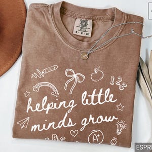 May include: A brown t-shirt with white text that reads "helping little minds grow". The shirt is decorated with drawings of a rainbow, a pencil, a lightbulb, a bow, an apple, and the numbers 1, 2, and 3.