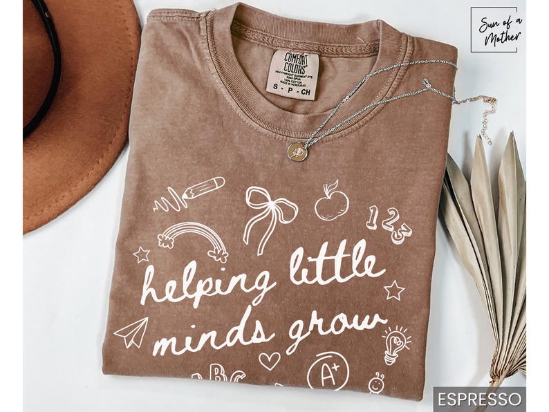May include: A brown t-shirt with white text that reads "helping little minds grow". The shirt is decorated with drawings of a rainbow, a pencil, a lightbulb, a bow, an apple, and the numbers 1, 2, and 3.