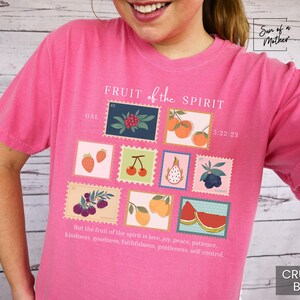 May include: Pink t-shirt with a graphic design of fruit in postage stamps. The text "FRUIT of the SPIRIT" is printed in white on the shirt. The scripture reference "GAL 5:22-23" is printed below the graphic. The text "But the fruit of the spirit is love, joy, peace, patience, kindness, goodness, faithfulness, gentleness, self control." is printed at the bottom of the shirt.