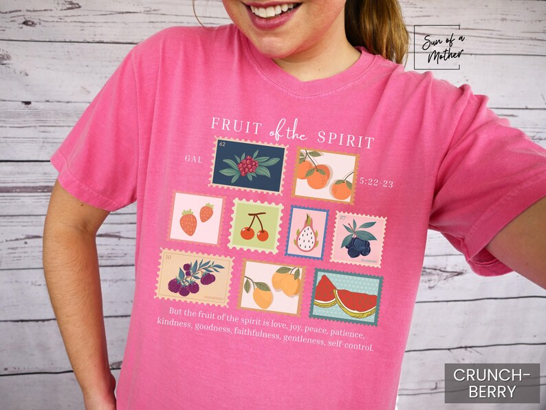 May include: Pink t-shirt with a graphic design of fruit in postage stamps. The text "FRUIT of the SPIRIT" is printed in white on the shirt. The scripture reference "GAL 5:22-23" is printed below the graphic. The text "But the fruit of the spirit is love, joy, peace, patience, kindness, goodness, faithfulness, gentleness, self control." is printed at the bottom of the shirt.