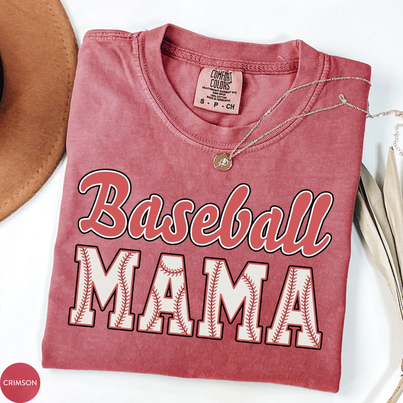 May include: A crimson red t-shirt with the text "Baseball Mama" in white and red lettering. The letters are outlined in black and have a baseball stitch design.
