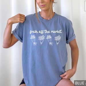 May include: A blue jean-colored t-shirt with the text "fresh off the market" above four white floral bouquets. The shirt has short sleeves and a relaxed fit. The text and floral design are in white.