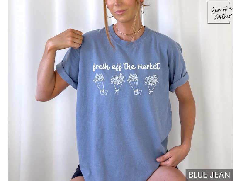 May include: A blue jean-colored t-shirt with the text "fresh off the market" above four white floral bouquets. The shirt has short sleeves and a relaxed fit. The text and floral design are in white.