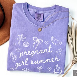May include: A violet Comfort Colors t-shirt with the phrase "pregnant girl summer" in white script, along with summer-themed graphics. A gold necklace and a brown hat are also visible.