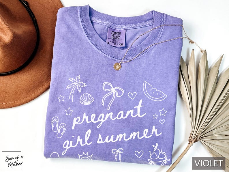 May include: A violet Comfort Colors t-shirt with the phrase "pregnant girl summer" in white script, along with summer-themed graphics. A gold necklace and a brown hat are also visible.