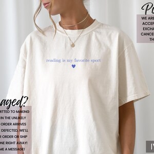 Puede incluir: Camiseta de color marfil con la frase "reading is my favorite sport" en azul claro, con un coraz&oacute;n azul. La camiseta tiene un corte holgado. El logotipo de la marca "Sun of a Mother" est&aacute; en la esquina superior izquierda.