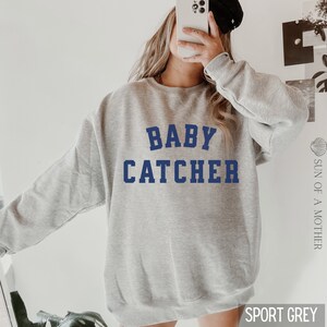 May include: A gray sweatshirt with the text "BABY CATCHER" in blue lettering. The sweatshirt is a relaxed fit and has a crew neck.