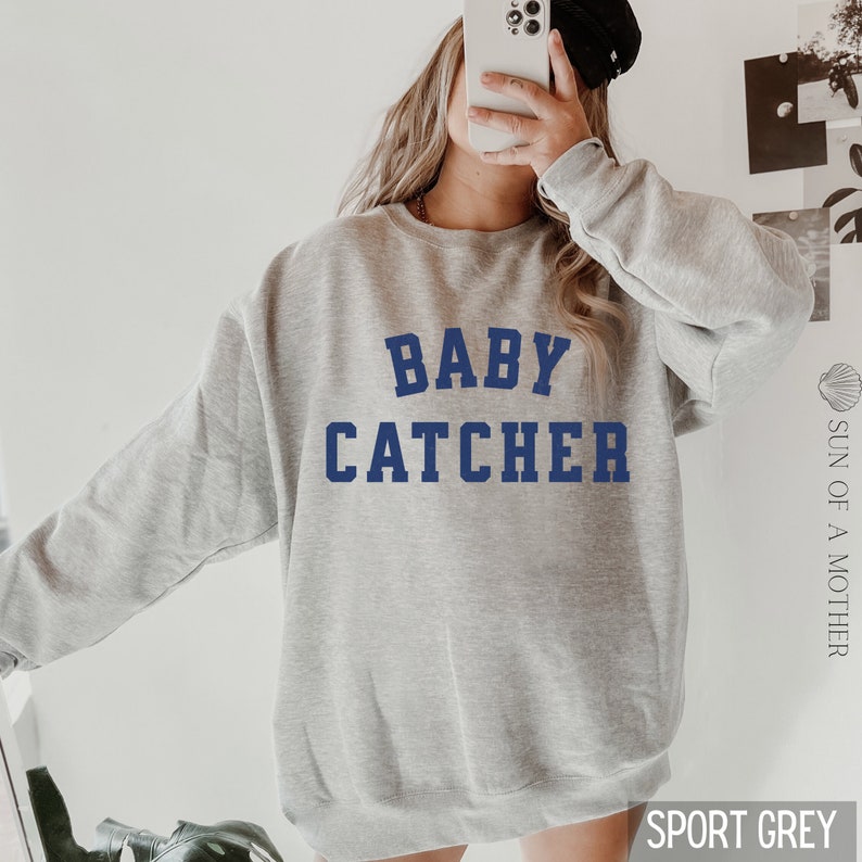 May include: A gray sweatshirt with the text "BABY CATCHER" in blue lettering. The sweatshirt is a relaxed fit and has a crew neck.
