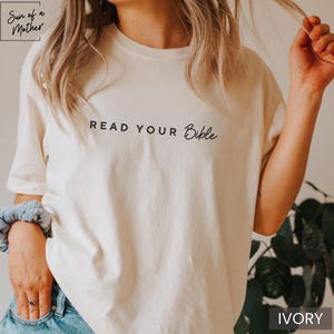 May include: A white t-shirt with the text "Read Your Bible" in black lettering. The shirt is shown on a model and the background is a blurry image of green plants. The color of the shirt is ivory.