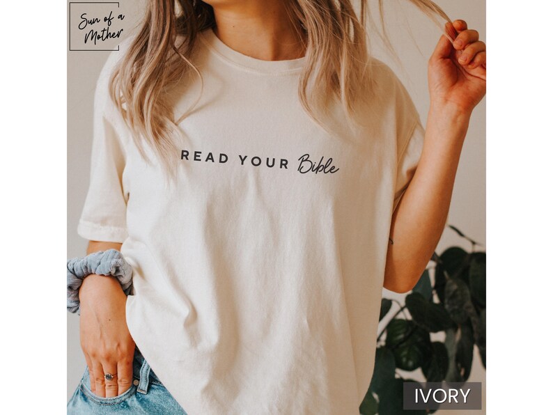 May include: A white t-shirt with the text "Read Your Bible" in black lettering. The shirt is shown on a model and the background is a blurry image of green plants. The color of the shirt is ivory.