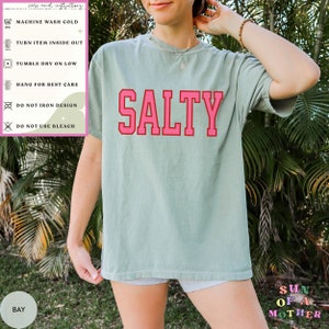 May include: A light green t-shirt with the word "SALTY" printed in pink letters with a red outline. The shirt is being worn by a person. The care instructions are listed on the left side of the image. The text "SUN OF A MOTHER" is printed in colorful letters on the bottom right corner of the image.