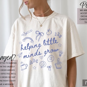 May include: White t-shirt with a blue graphic that says "helping little minds grow". The graphic includes illustrations of a rainbow, an apple, a pencil, a lightbulb, a book, a coffee cup, a bow, and the letters "ABC". The shirt is available in ivory.