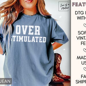May include: A blue jean t-shirt with the text "OVER STIMULATED" printed in white. The shirt is a relaxed fit and has a soft, vintage feel.