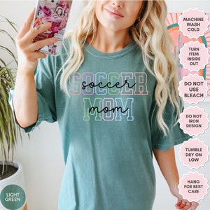 May include: A light green t-shirt with the text "soccer mom" in rainbow colors. The text is outlined in black. The shirt has a crew neck and short sleeves.