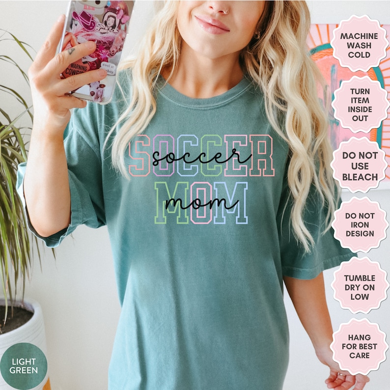 May include: A light green t-shirt with the text "soccer mom" in rainbow colors. The text is outlined in black. The shirt has a crew neck and short sleeves.