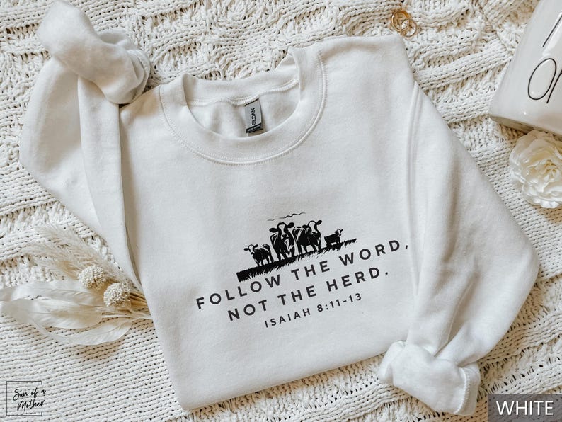 May include: White crewneck sweatshirt with the text "FOLLOW THE WORD, NOT THE HERD" and the Bible verse Isaiah 8:11-13. A black graphic of cows is above the text. The sweatshirt is on a white textured surface.