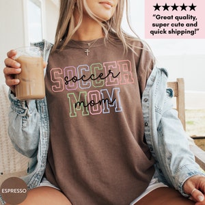 May include: A brown t-shirt with the text "SOCCER MOM" in rainbow colors. The text is outlined in black. The shirt is being worn by a person.