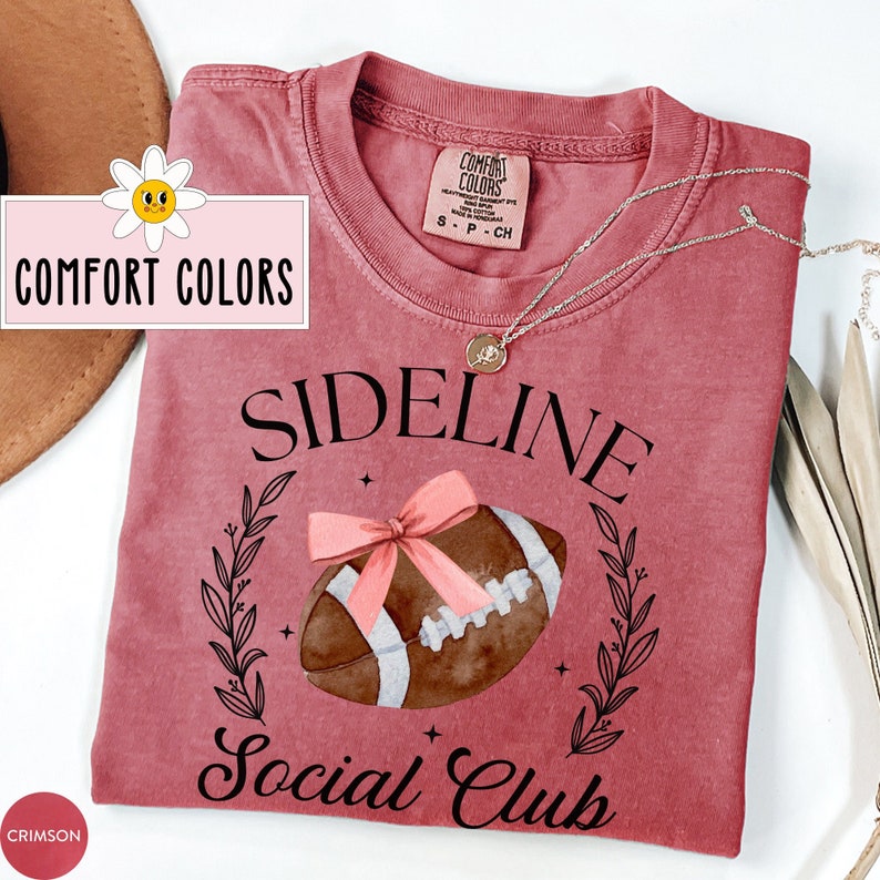 以下が含まれることがあります： クリムゾンレッドのTシャツに、水彩画のフットボールのデザインと「Sideline Social Club」の文字がプリントされています。