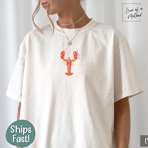 Może przedstawiać: Biały t-shirt z grafiką czerwonego homara i tekstem "I'm catching" w kolorze niebieskim. Koszulka wykonana jest z miękkiego, wygodnego materiału.