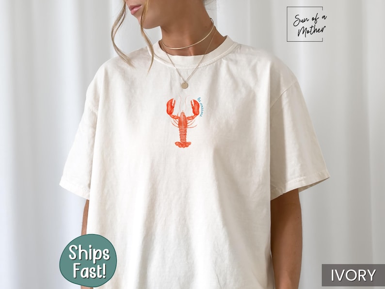 Może przedstawiać: Biały t-shirt z grafiką czerwonego homara i tekstem "I'm catching" w kolorze niebieskim. Koszulka wykonana jest z miękkiego, wygodnego materiału.