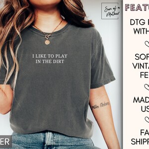May include: A dark gray t-shirt with the text "I like to play in the dirt" printed on the front. The shirt is made with a soft, vintage feel and is printed with ink using a direct-to-garment printing method.