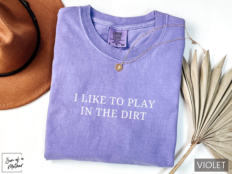 May include: A purple t-shirt with the text "I like to play in the dirt" printed in white. The shirt is made of a soft, comfortable material and has a relaxed fit.