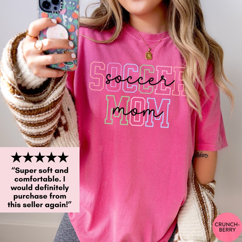 May include: Pink t-shirt with the text "soccer mom" in rainbow colors. The text is outlined in white. The shirt is being held by a person.
