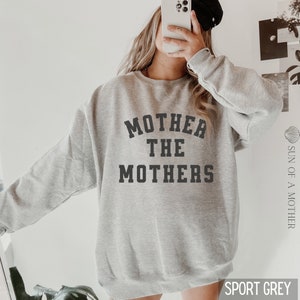 K&ouml;nnte beinhalten: Ein grauer Pullover mit dem Aufdruck "Mother the Mothers" in Schwarz auf der Vorderseite. Der Pullover ist locker geschnitten und hat einen Rundhalsausschnitt.