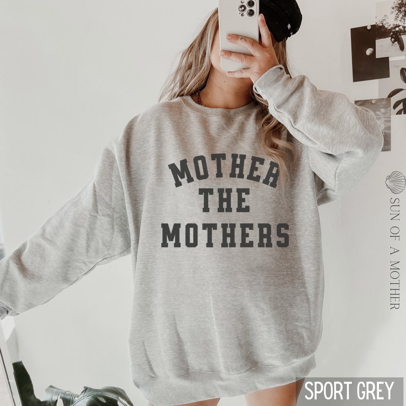 K&ouml;nnte beinhalten: Ein grauer Pullover mit dem Aufdruck "Mother the Mothers" in Schwarz auf der Vorderseite. Der Pullover ist locker geschnitten und hat einen Rundhalsausschnitt.