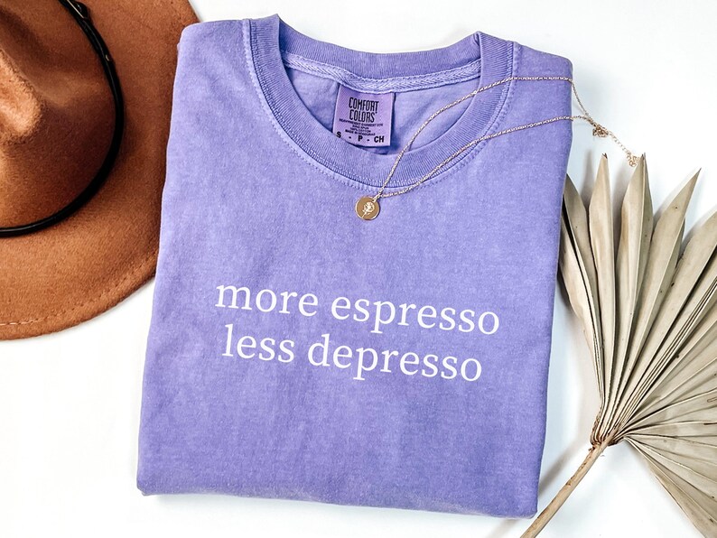 Op de afbeelding: Een lavendelkleurig Comfort Colors T-shirt met de witte tekst "more espresso less depresso". Een gouden ketting en een bruine hoed zijn ook in de afbeelding te zien. Het shirt is opgevouwen en heeft een vintage look.