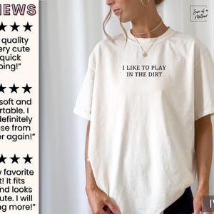 May include: A white t-shirt with the text "I like to play in the dirt" printed on the front. The shirt is shown on a model and the color is ivory.