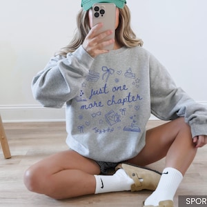 May include: Sport grey sweatshirt with the text "just one more chapter" in blue. The design includes book-themed illustrations. The sweatshirt is a comfortable, casual style.