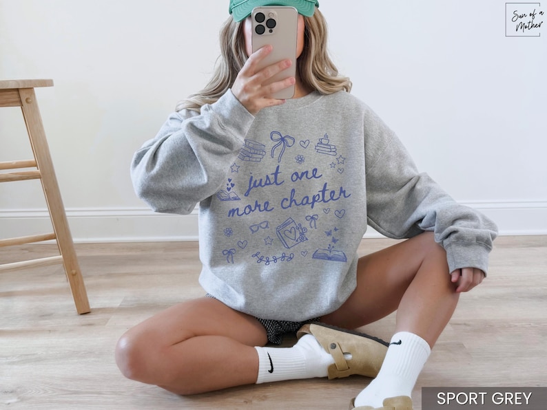 May include: Sport grey sweatshirt with the text "just one more chapter" in blue. The design includes book-themed illustrations. The sweatshirt is a comfortable, casual style.