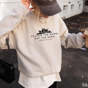 May include: Sand-colored sweatshirt with the text "FOLLOW THE WORD, NOT THE HERD" and a graphic of cows. The text is in black, with the Bible verse Isaiah 8:11-13 below. The sweatshirt is a casual style.