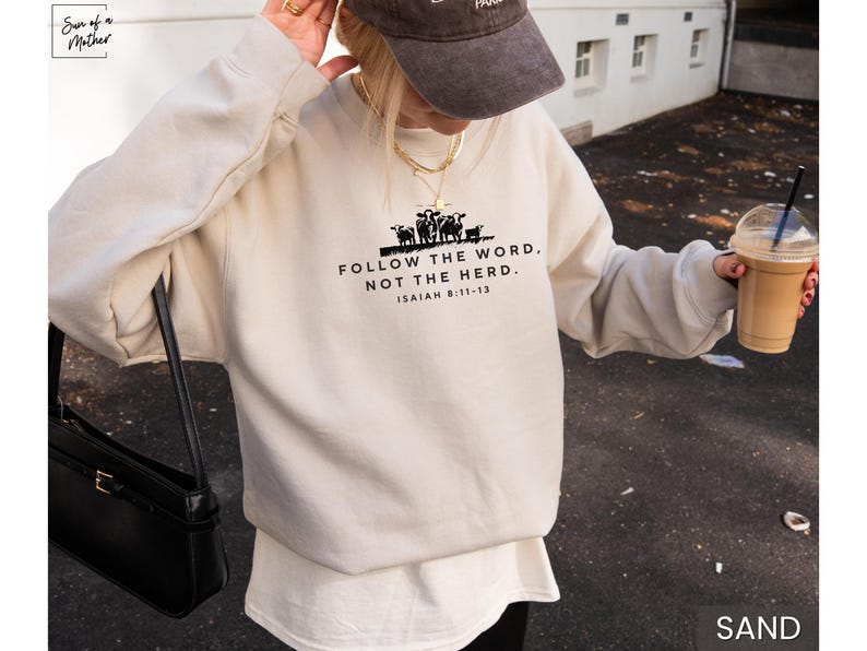 May include: Sand-colored sweatshirt with the text "FOLLOW THE WORD, NOT THE HERD" and a graphic of cows. The text is in black, with the Bible verse Isaiah 8:11-13 below. The sweatshirt is a casual style.