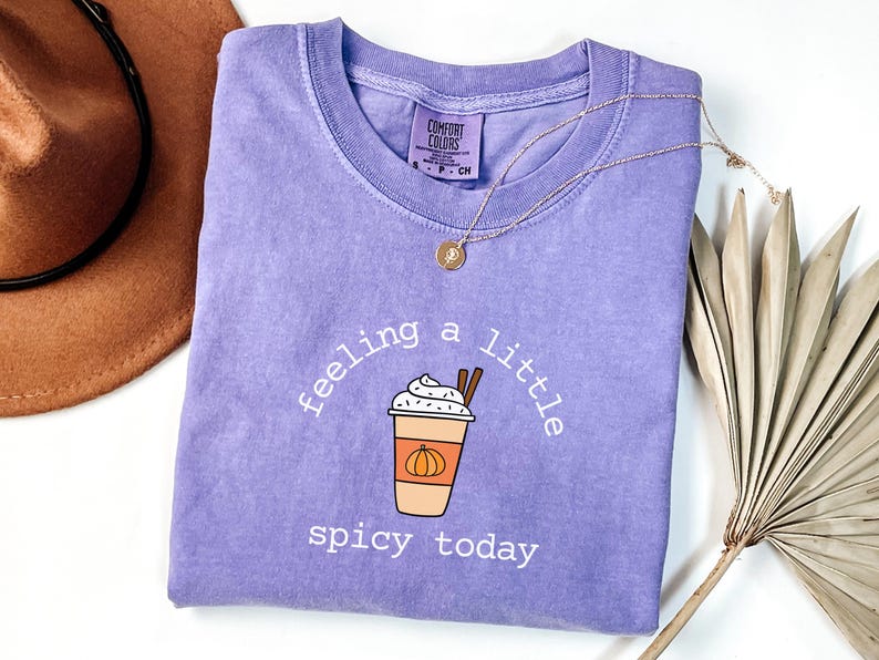 May include: A lilac-colored t-shirt with the phrase "feeling a little spicy today" and a graphic of a pumpkin spice latte. A gold necklace and a brown hat are also in the image.