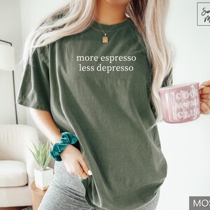 Op de afbeelding: Mosgroen T-shirt met de witte tekst "more espresso less depresso". Een roze mok met "Cool Mom Club" wordt in de hand gehouden. Het shirt heeft een casual stijl. Er is ook een turkooiskleurige scrunchie te zien.