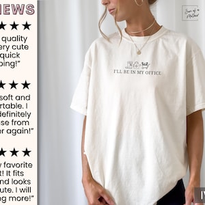 May include: A white t-shirt with the text "I'll be in my office" and a graphic of a coffee cup, watering can, and a wheelbarrow. The shirt is available in ivory.