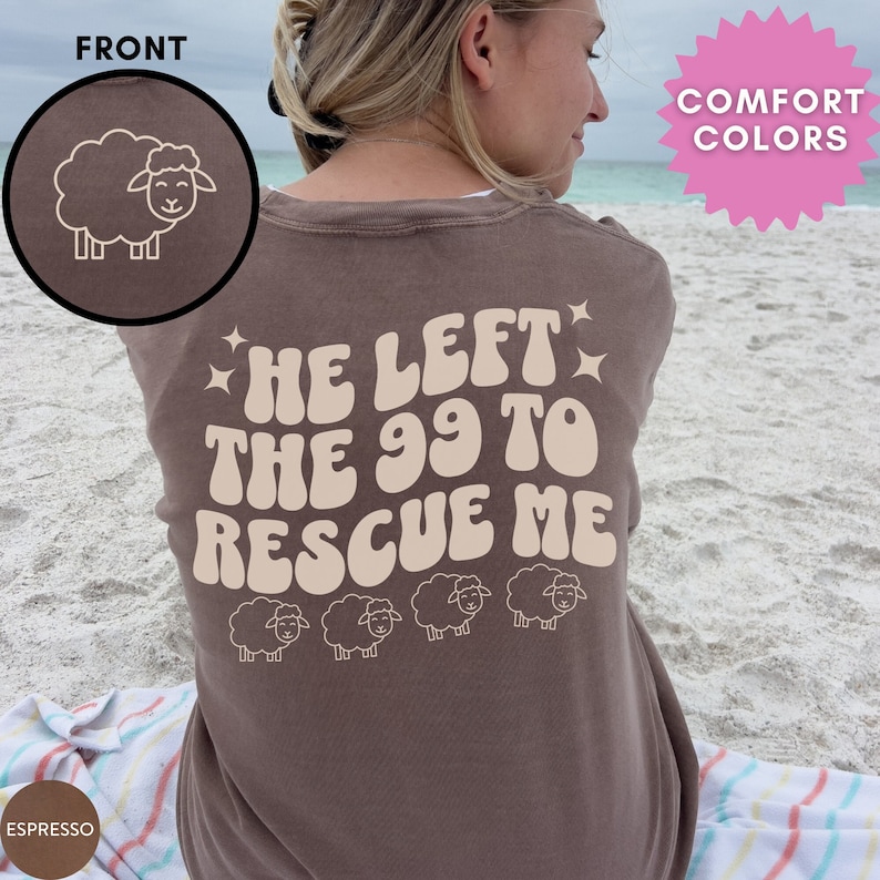 May include: A brown t-shirt with the text "He left the 99 to rescue me" in a retro font. The text is in a light beige color. There are five small sheep silhouettes below the text. The shirt is being worn by a person standing on a beach.