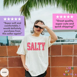May include: A white t-shirt with the word "SALTY" printed in pink letters. The shirt is being worn by a person with sunglasses. The background is a brick wall with a palm tree and a building in the distance.